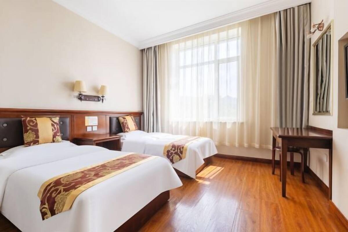 Habitació d'hotel amb dos llits, un terra de fusta, un escriptori i una finestra amb cortines.