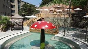 Zona de piscina con una fuente en forma de hongo, sillas de descanso, sombrillas y una escalera que conduce a una terraza.