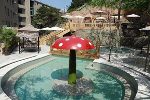 A pool area with a mushroom-shaped water feature, lounge chairs, umbrellas, and a staircase leading to a terrace.