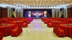Un salón de banquetes con mesas y sillas redondas, un escenario con una pantalla grande e iluminación empotrada en el techo.