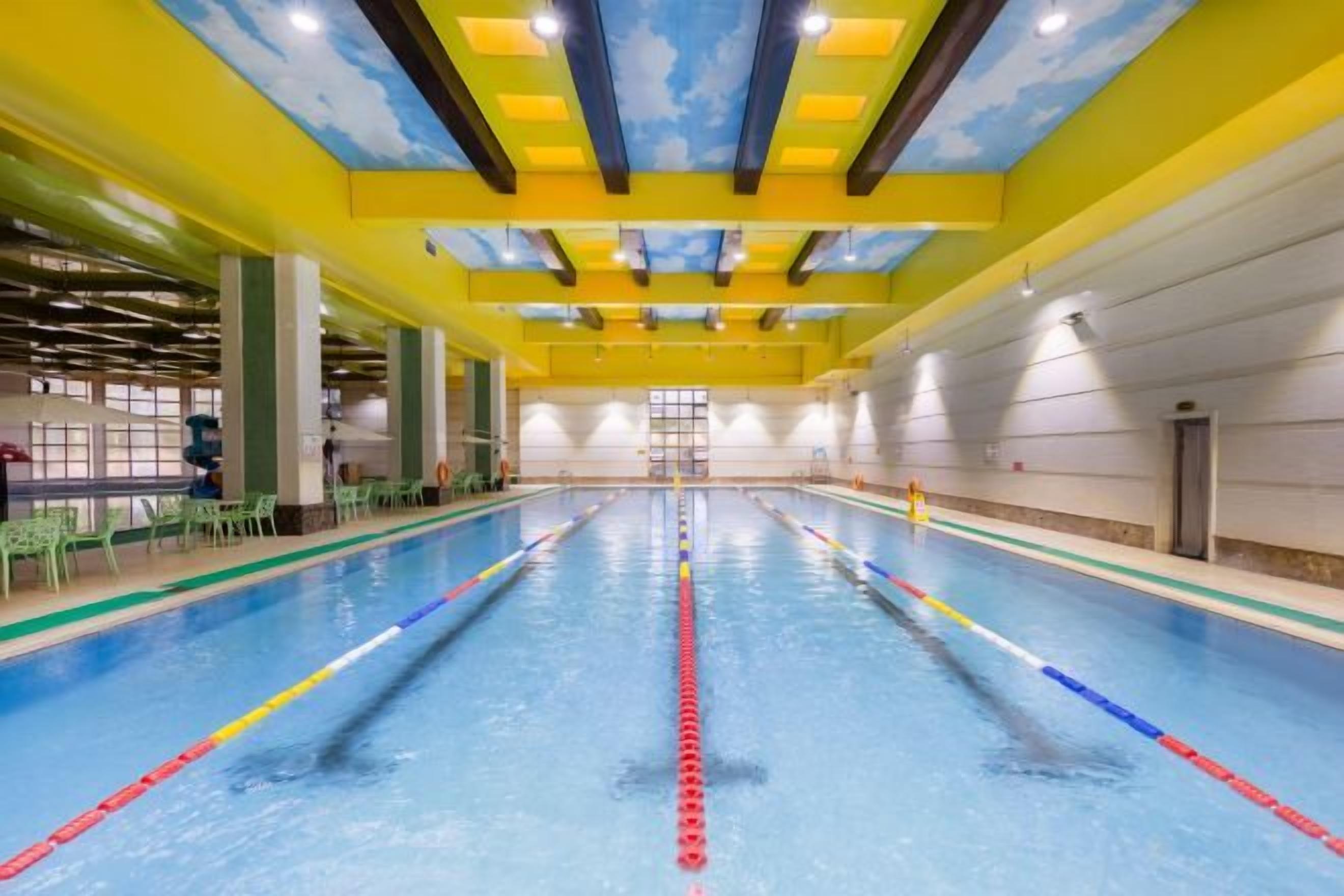 Una piscina interior amb separadors de carrils ben definits, un sostre amb disseny blau i groc, i un ambient clar i ben il·luminat.