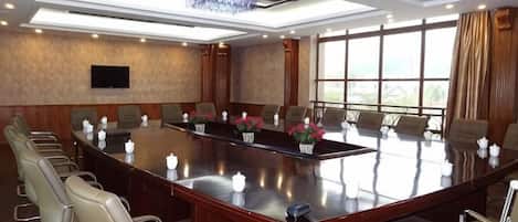 Un salón de conferencias con una larga mesa, varias sillas, un televisor de pantalla plana y ventanas amplias.