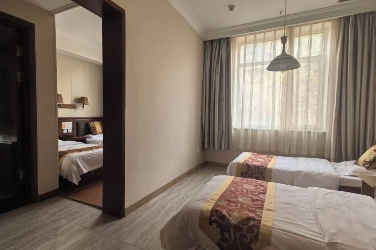 Habitació d'hotel amb dos llits, una finestra gran amb cortines, una làmpada penjant i una porta visible que condueix a una altra habitació.