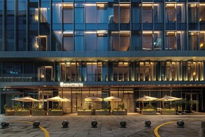 Un hotel moderno con fachada de vidrio, asientos exteriores con sombrillas y una cafetería visible.