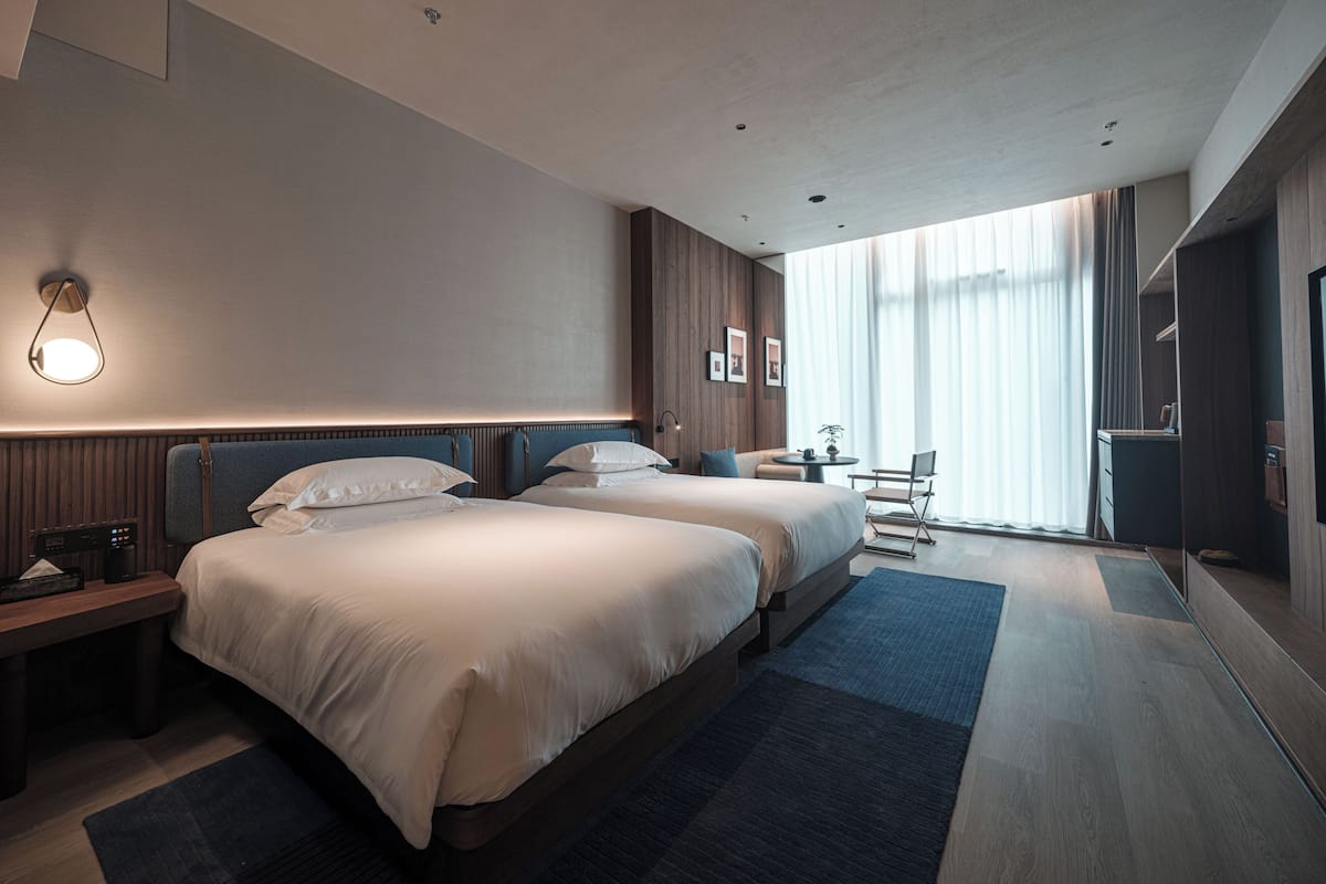 A modern hotel room with two beds, a desk, a chair, a small table, and a large window with curtains.