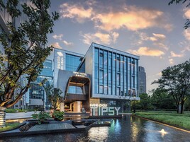 A modern hotel with a glass facade, a water feature in front, and signage indicating 'TIPSY HOTEL' and 'HANG ZHOU'.