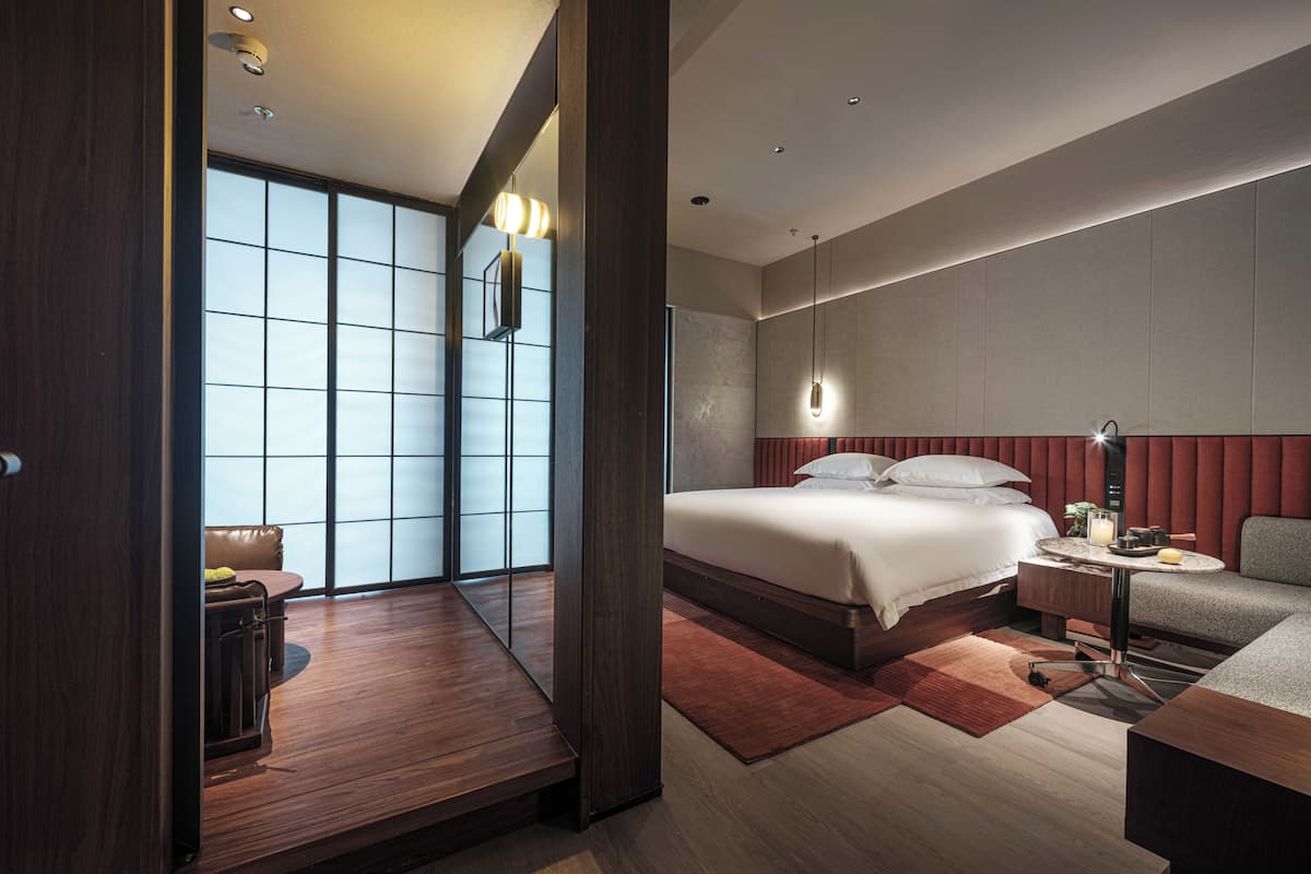 A modern hotel room with a large bed, a desk, a chair, and a view through a large window.