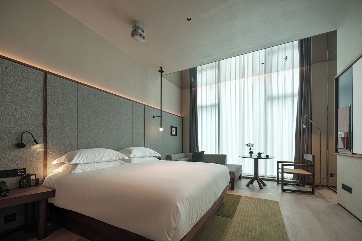 A modern hotel room with a large bed, a desk, a chair, and a small table.