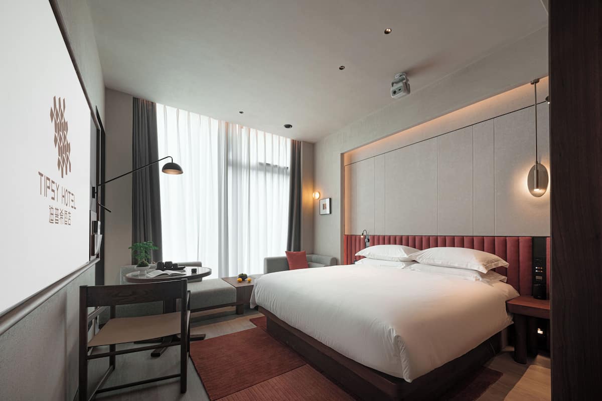 A hotel room with a large bed, bedside tables, a desk with a chair, a window with curtains, and a wall-mounted TV.