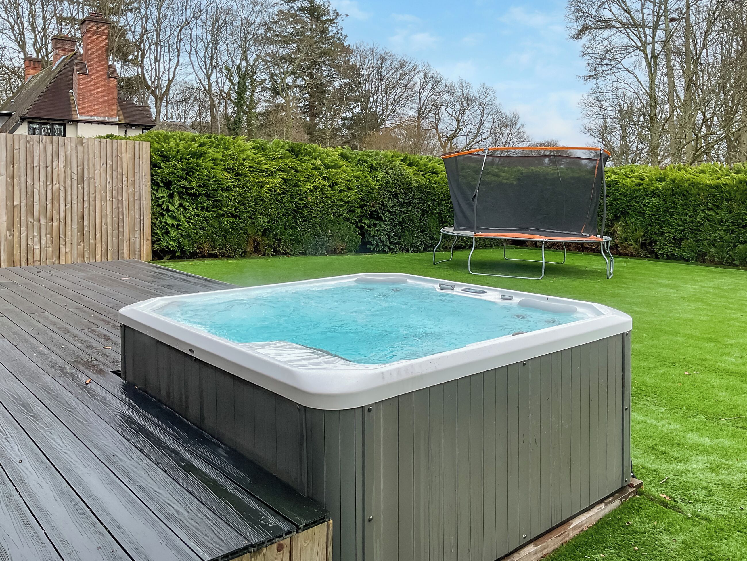 Un jacuzzi en un patio trasero con una terraza de madera, un trampolín y un seto.