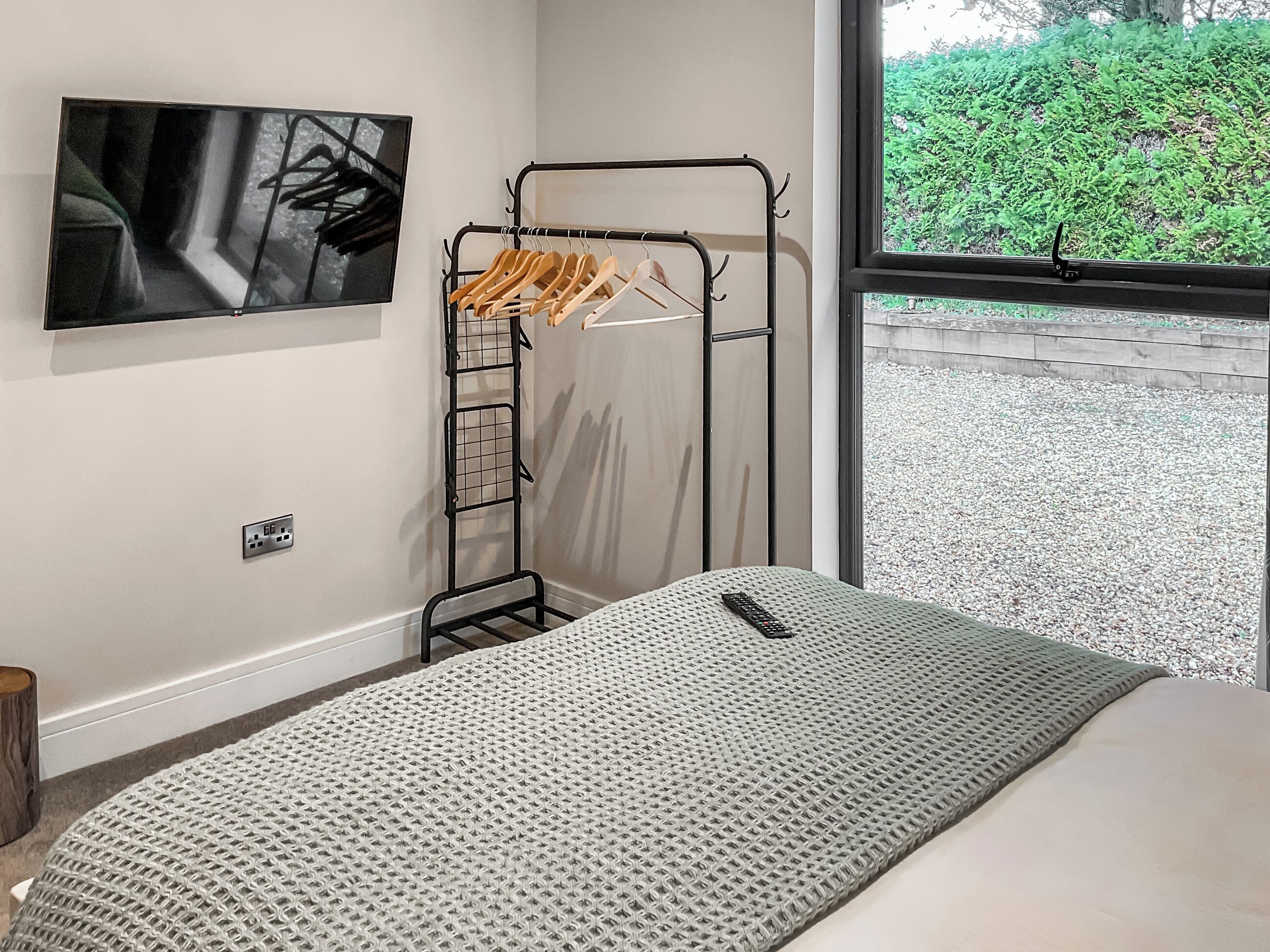 A bedroom with a bed, a wall-mounted TV, a clothes drying rack, and a window with a view of greenery.