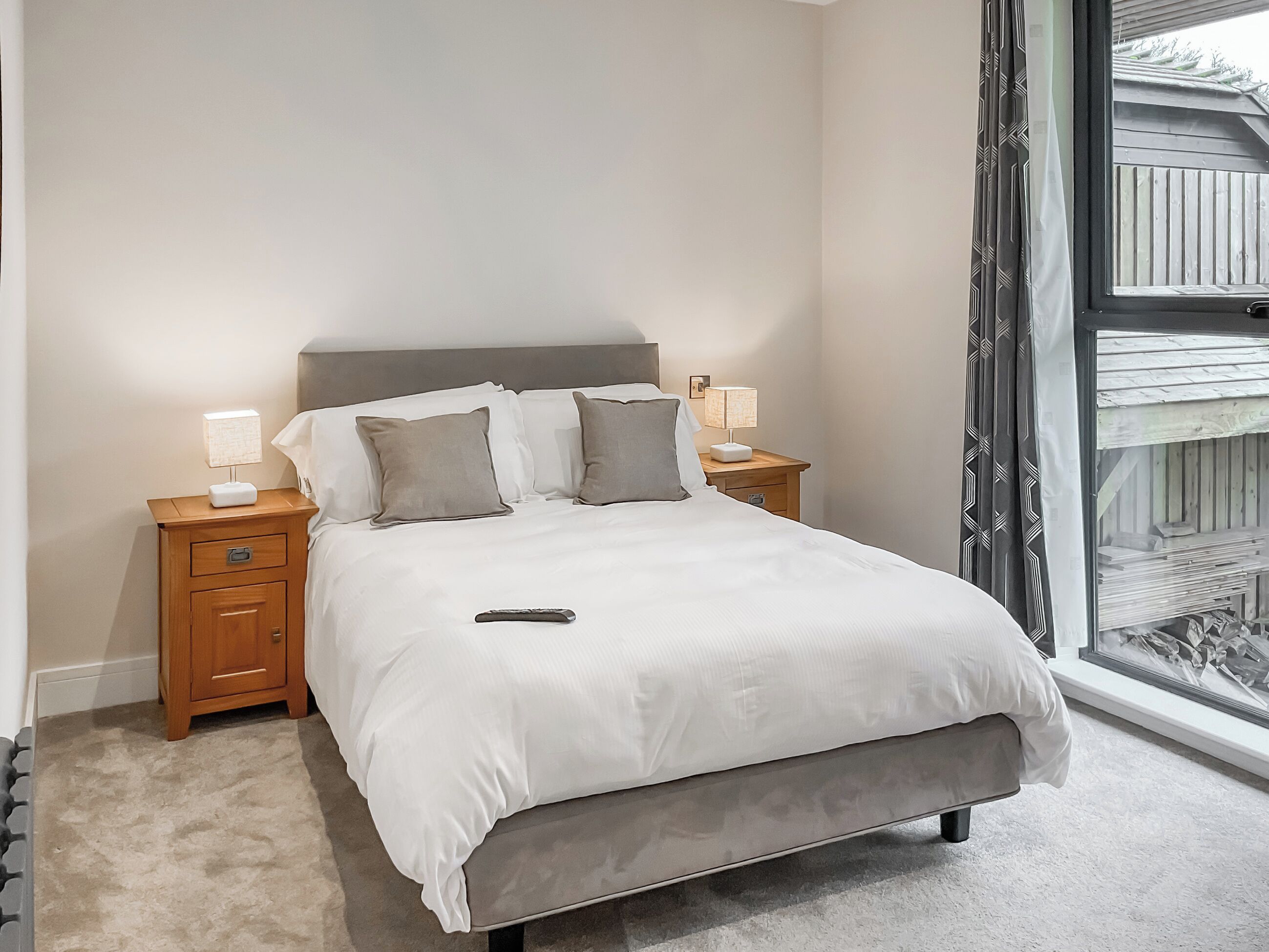 A neatly made bed with a grey headboard, two bedside tables with lamps, and a window with a view of a wooden deck.