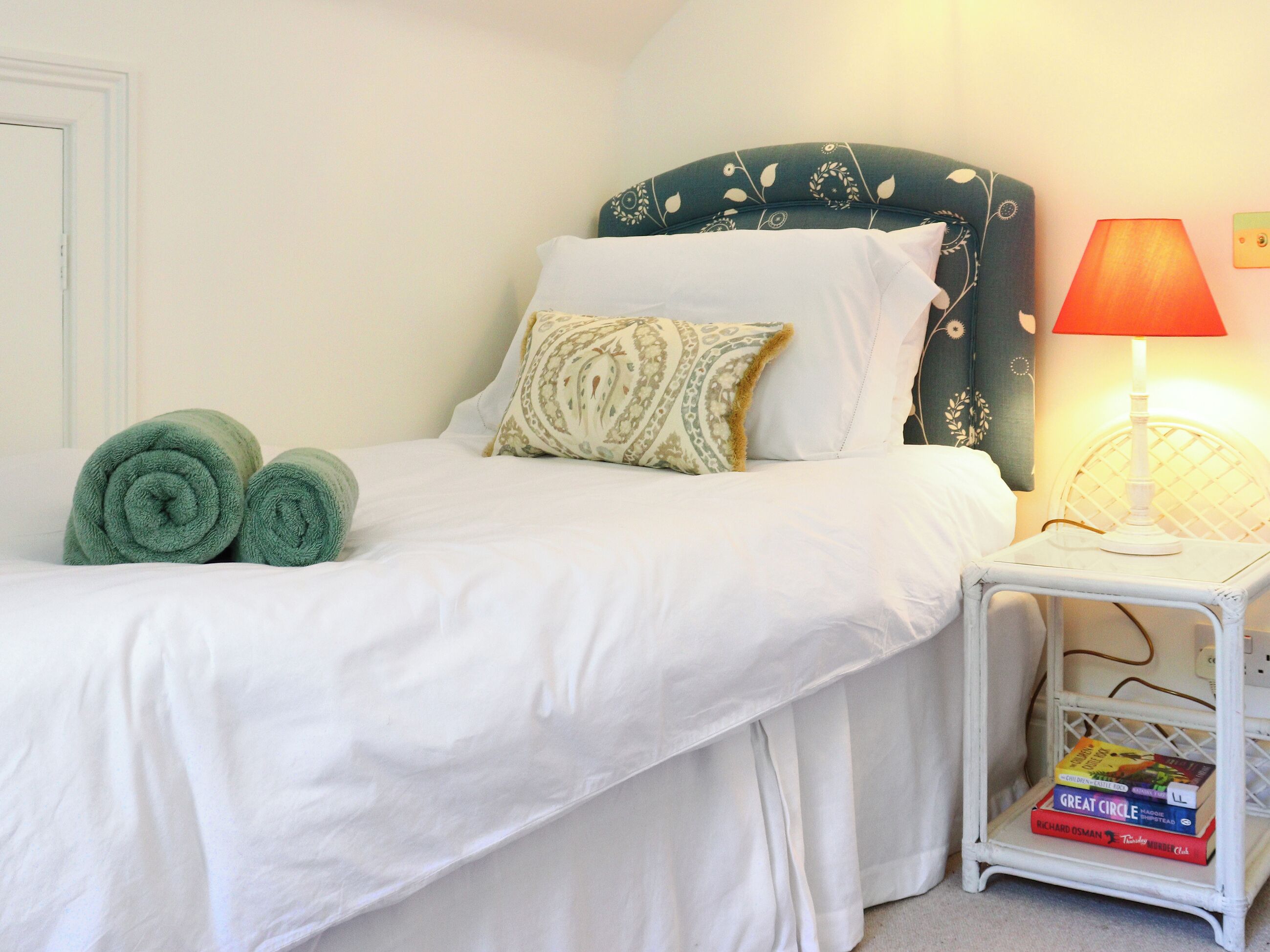 A neatly made bed with white linens, a green headboard, and a bedside table with a lamp and a book shelf.