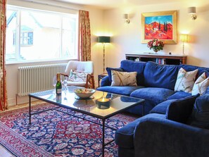 A living room with a blue sofa, a glass coffee table, a white armchair, a patterned rug, a painting on the wall, and a radiator. - 3 bedroom accommodation in Near Bath (Bradford-on-Avon)