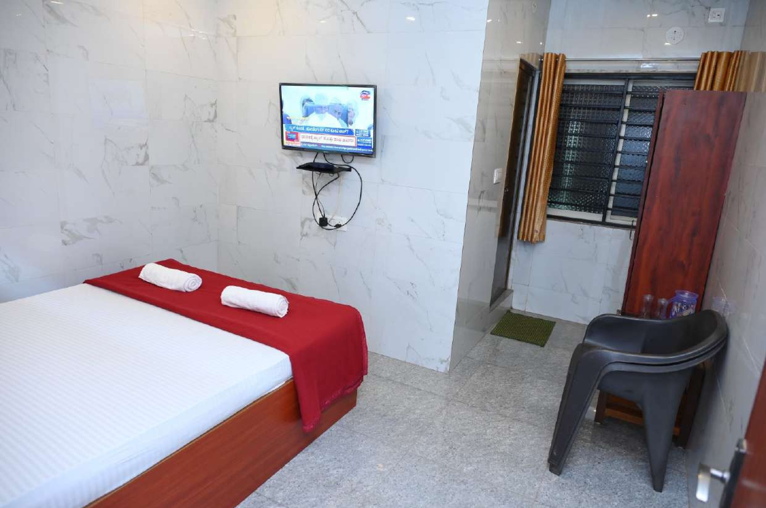 Una camera d'albergo con un letto, una TV a muro, una finestra con tende e un piccolo sgabello.