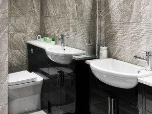 Et moderne badeværelse med en hvid vask, toilet og bidet. Væggene er beklædt med fliser i et marmorlignende mønster.