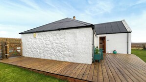 A white building with a dark roof, a wooden deck, and a grassy yard. - 1 bedroom accommodation in Portree (Portree)