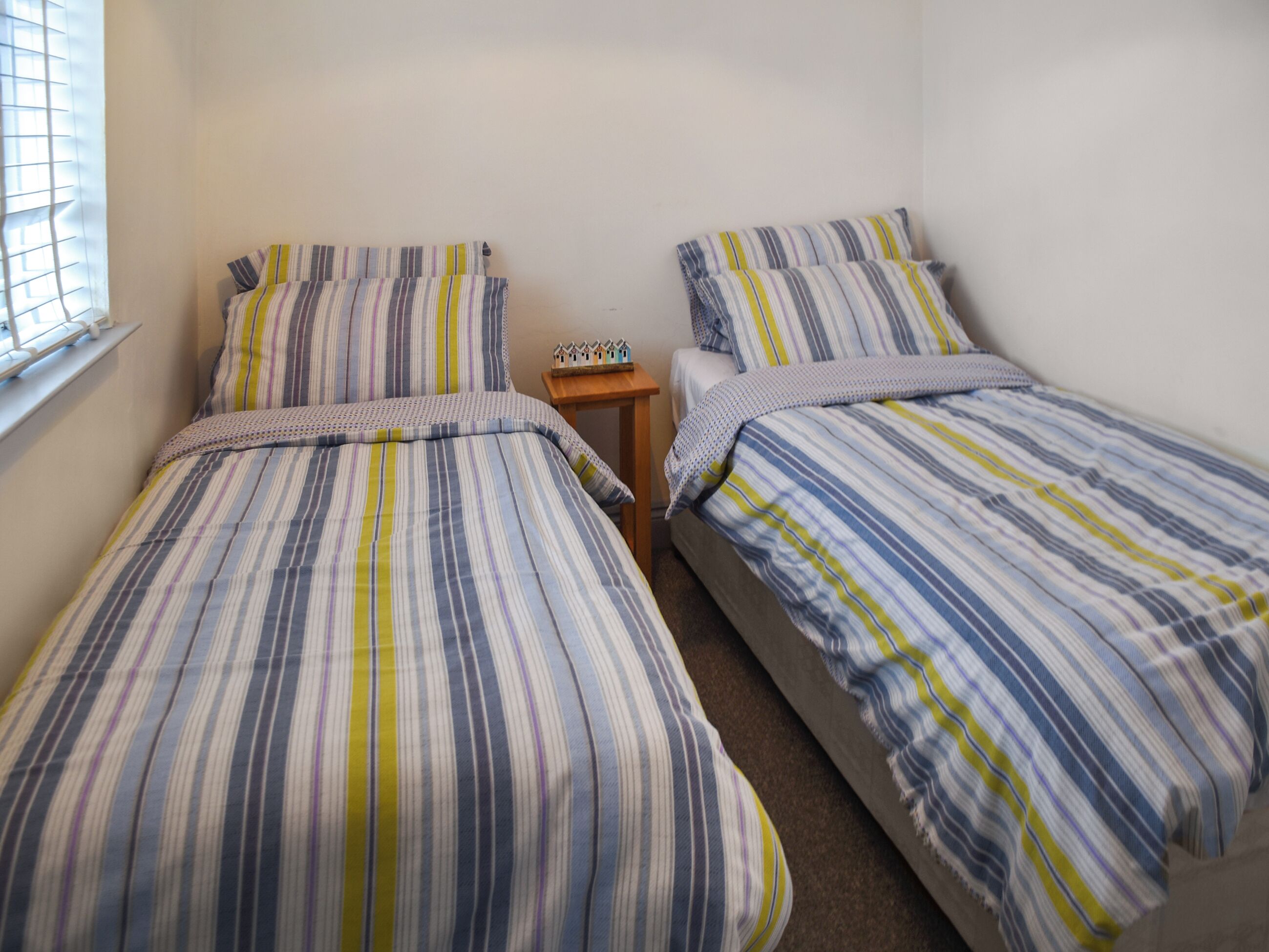 A room with two beds, each with striped bedding, a small wooden nightstand, and a window with blinds.