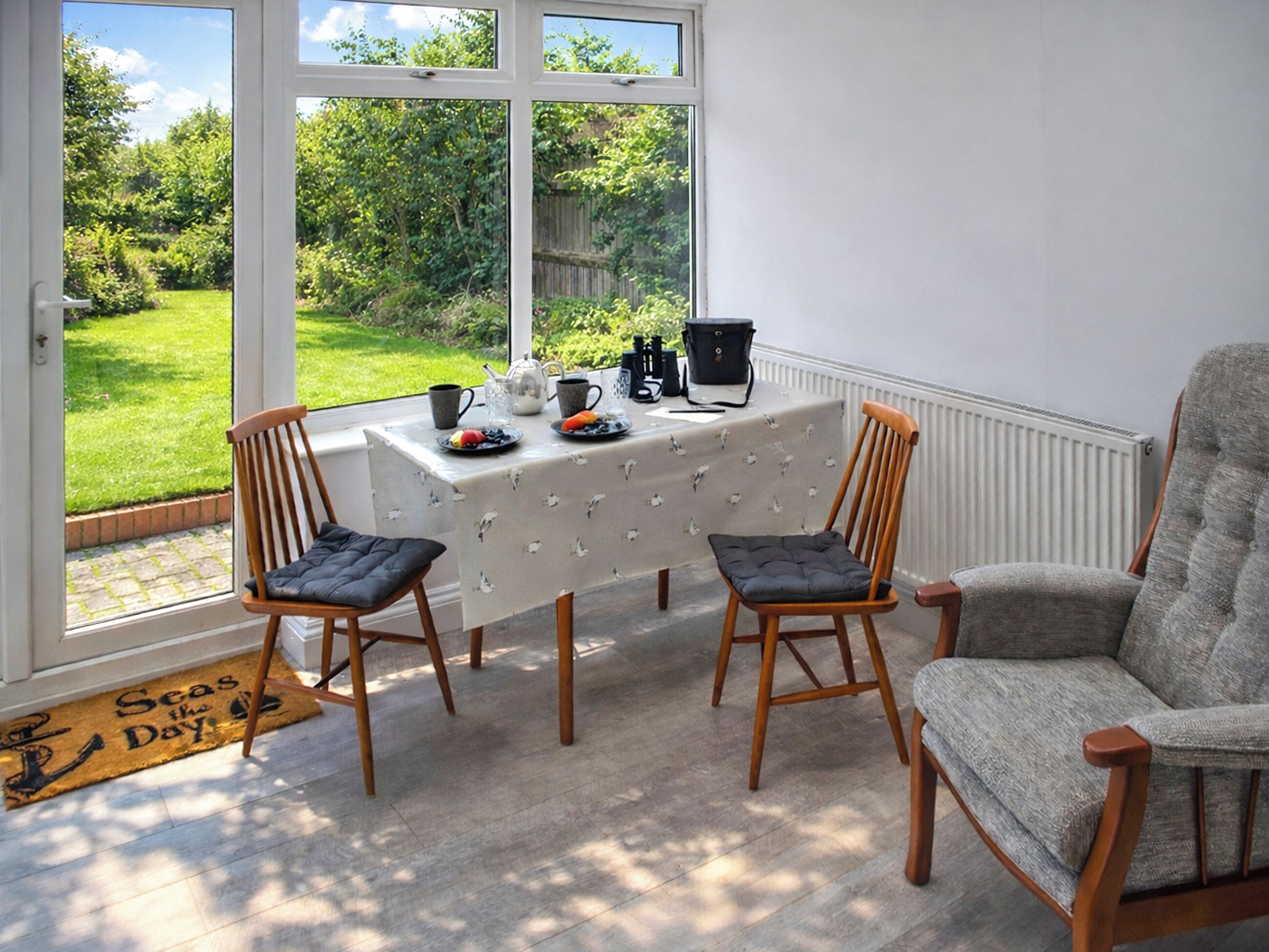 A room with a table set for two, a chair, a radiator, and a view of a garden through large windows.