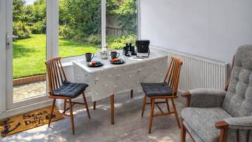 A room with a table set for two, a chair, a radiator, and a view of a garden through large windows.