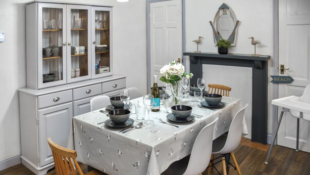 Un coin repas avec une table dressée pour quatre personnes, entouré de chaises en bois, d’un buffet aux portes vitrées et d’une cheminée surmontée d’un miroir.