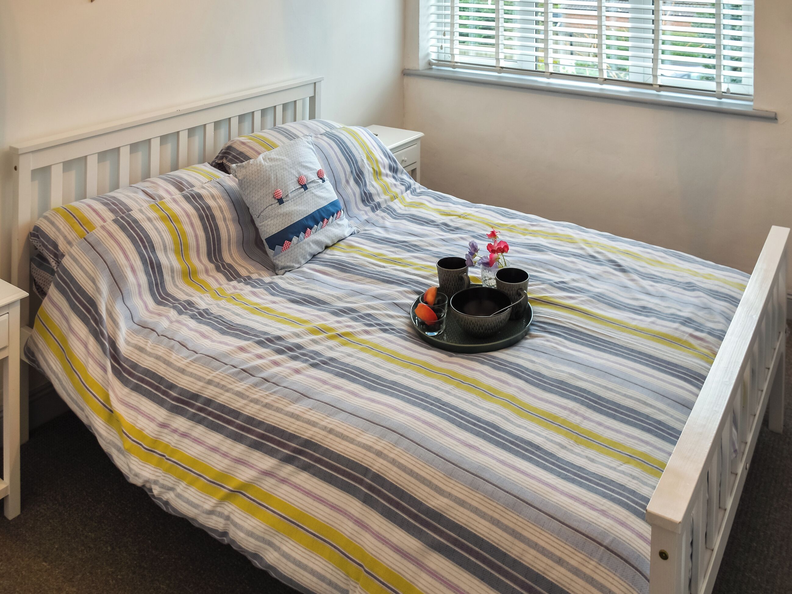 A bed with striped bedding, a bedside table, and a window with blinds.