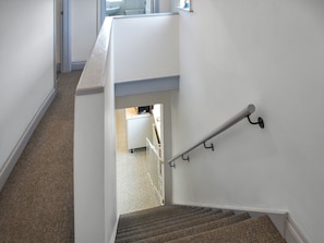 Un escalier avec une rampe, des marches recouvertes de moquette et une vue sur une pièce comprenant un coin cuisine.
