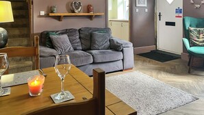 A living room with a grey sofa, a wooden coffee table with a candle and wine glass, a 'Be our guest' sign, and a door with a fire alarm sign. - 1 bedroom accommodation in Kelbrook (Kelbrook)