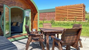 An outdoor seating area with a wooden table and chairs, a green archway entrance, and a wooden building in the background. - Chesnut Pod - UK51519 (Newton Stewart)