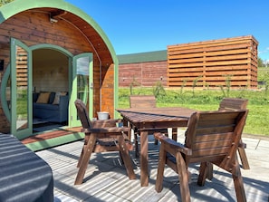 An outdoor seating area with a wooden table and chairs, a green archway entrance, and a wooden building in the background. - Chesnut Pod - UK51519 (Newton Stewart)