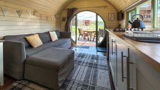 A cozy cabin interior with a sofa, ottoman, wooden ceiling, and a view through a door to an outdoor patio. - Chesnut Pod - UK51519 (Newton Stewart)
