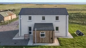 A two-story building with a flat roof, a small attached wooden structure, and a paved driveway. - Waking up to the wonderful views of the wide beaches and open skies. (North Uist)