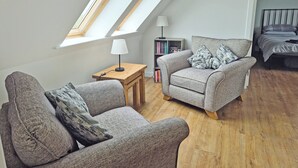 A loft bedroom with a skylight, a sofa, a side table, a lamp, a bookshelf, and a bed. - Waking up to the wonderful views of the wide beaches and open skies. (North Uist)