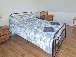 Ein ordentlich bezogenes Bett mit einem blumigen Überwurf, ein Nachttisch mit Schubladen, ein Stuhl und eine Lampe.