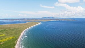 An aerial view of a coastal landscape with a beach, green fields, and a clear blue sky. - Waking up to the wonderful views of the wide beaches and open skies. (North Uist)