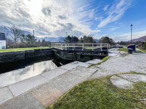 Un éclusier avec un pont piétonnier, un sentier pavé de pierres et une zone herbeuse, le tout sur fond de montagnes.