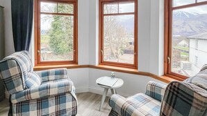 A corner room with two plaid chairs, a small table, and large windows with a view of a garden and mountains. - Flat 7, Canalside Apartments (Banavie, Fort William)