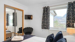 A bedroom with a bed, a chair, a mirror, a television, and a window with curtains. - Flat 7, Canalside Apartments (Banavie, Fort William)