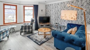 A living room with a blue sofa, a plaid armchair, a television, a coffee table, and a view through large windows. - Flat 7, Canalside Apartments (Banavie, Fort William)