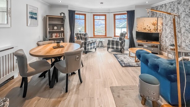 A modern living room with a round wooden table, grey upholstered chairs, a blue sofa, a television, and a bookshelf. - Flat 7, Canalside Apartments (Banavie, Fort William)