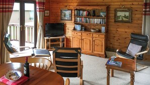 A cozy cabin interior with a wooden floor, a dining table with chairs, a television, a bookshelf, and a window with curtains. - The openness of the park and the closeness to the shore. (Acharacle, near Ardnamurchan)