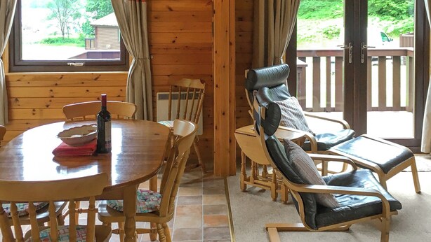 A wooden interior with a dining table, chairs, a reclining chair, a clock, and a view of greenery outside. - The openness of the park and the closeness to the shore. (Acharacle, near Ardnamurchan)