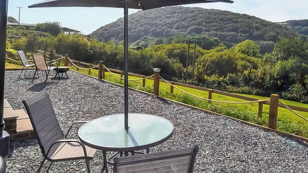 An outdoor patio with a table and chairs, a large umbrella, and a view of a mountainous landscape. - 1 bedroom accommodation in Ilfracombe (Ilfracombe)