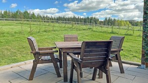 Eine Terrasse im Freien mit einem Holztisch und -stühlen, Blick auf ein saftiges Grasfeld und einen klaren Himmel.