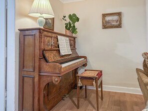Un piano de cua amb partitura, una taula petita amb un coixí, una planta en test i una fotografia encadrada a la paret.
