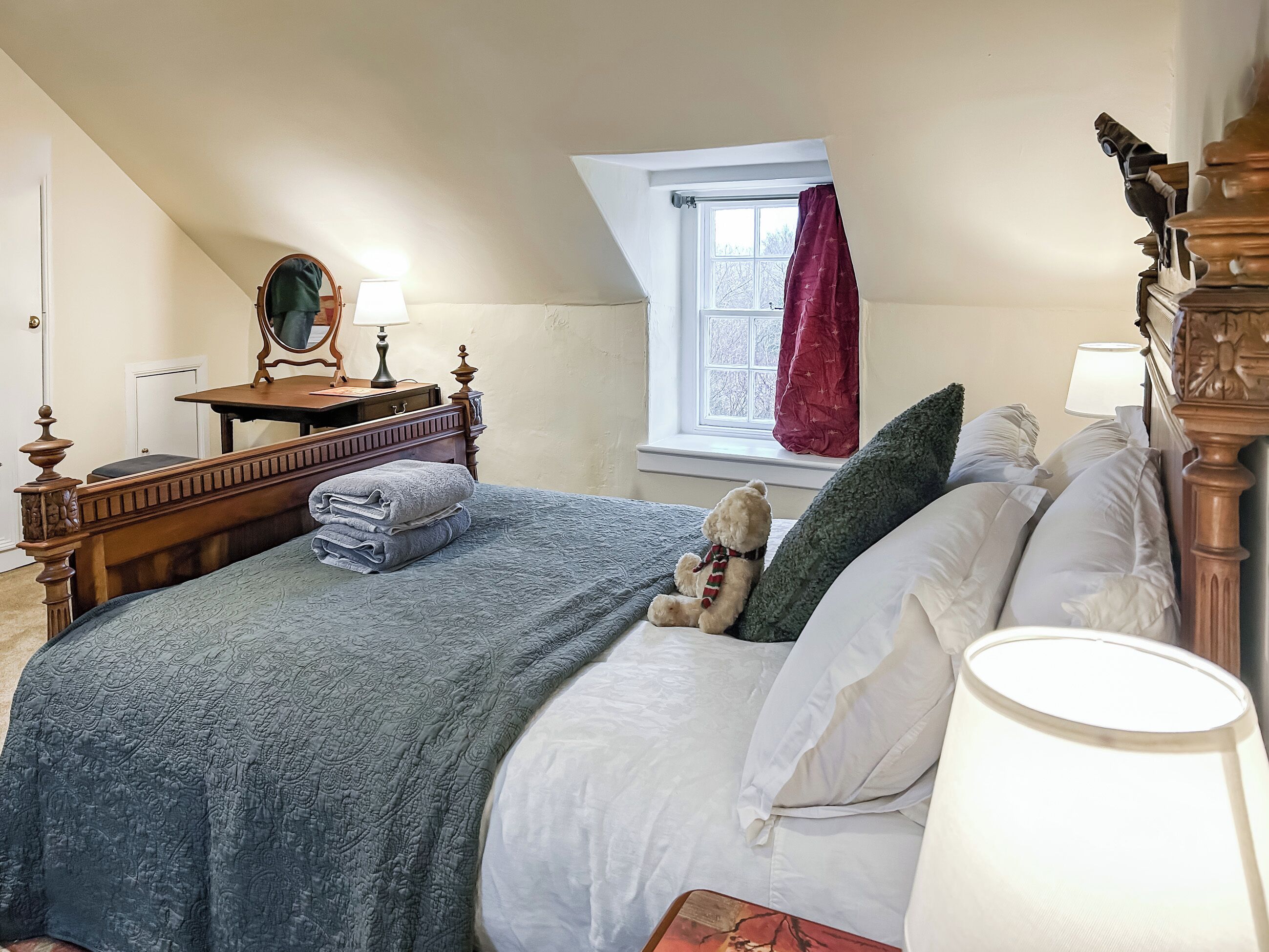 A bedroom with a bed, pillows, a teddy bear, a lamp, a dresser, a mirror, and a window with curtains.