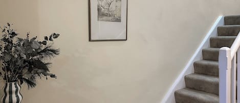 Un escalier avec des marches recouvertes de moquette et une rampe blanche, un tableau encadré au mur, et un vase rayé orné de feuillage, posé sur une table.