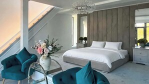 Quarto moderno com uma cama grande, duas poltronas verde-água, uma mesa de centro de vidro, um lustre e vista para o exterior.