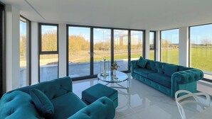 Uma sala de estar moderna com janelas amplas, um sofá verde-água, uma mesa de centro de vidro e uma cadeira branca.