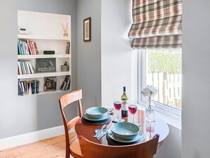 Ein Essbereich mit Tisch für zwei Personen, ein Bücherregal mit Büchern und Dekoration, und ein Fenster mit Blick auf Grünflächen.
